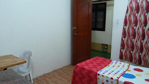 Una habitación pequeña con una mesa de madera, una silla blanca, una cama con colcha de lunares de colores y una cortina estampada.