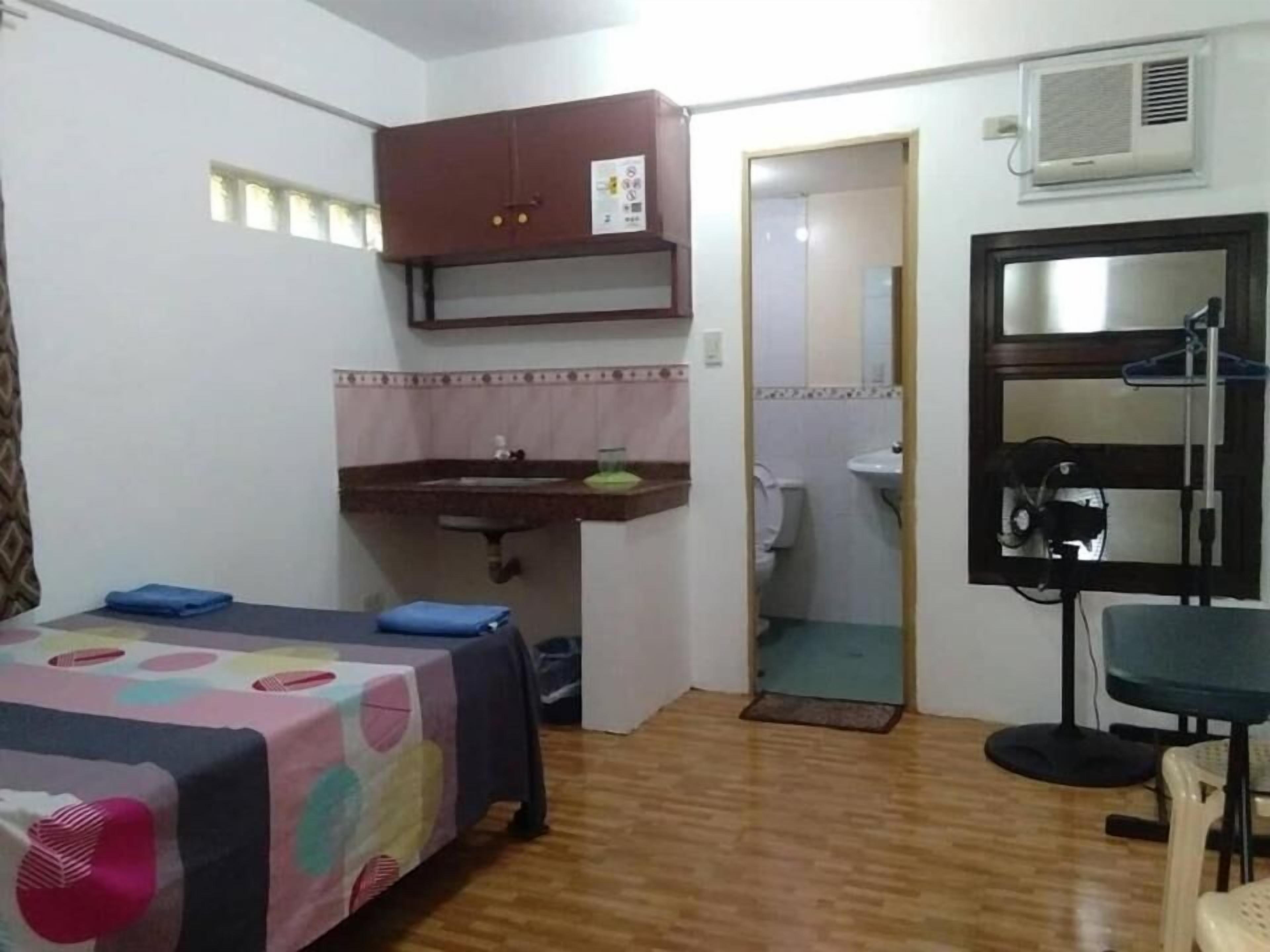 A small, compact room with a bed, a desk, a chair, a sink, and a mirror.