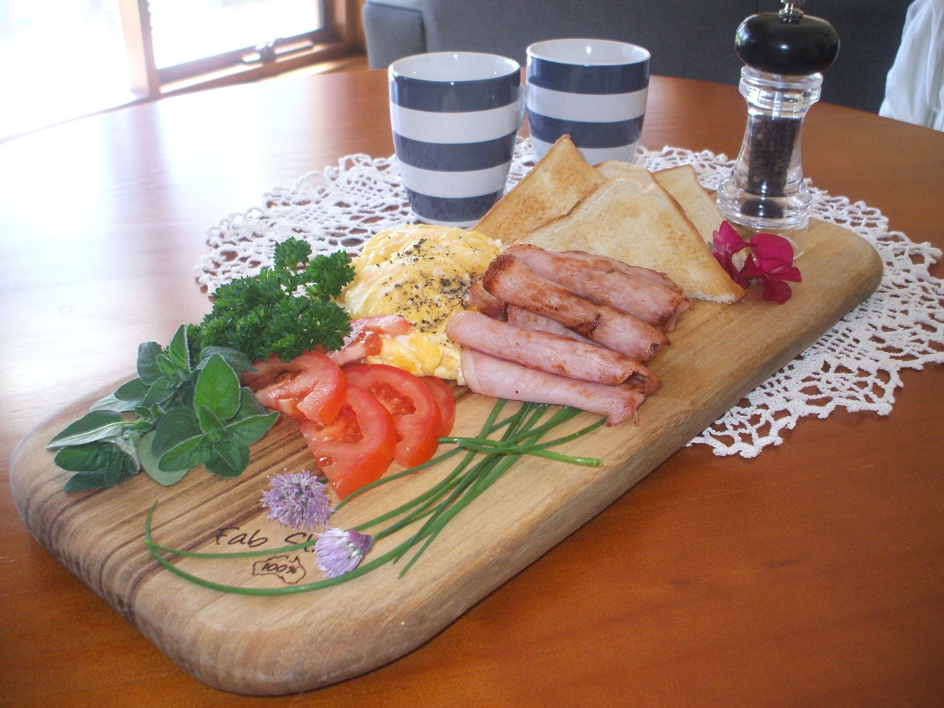 En træplanke med et udvalg af morgenmadsprodukter, herunder æg, bacon, tomater og krydderurter, placeret på et bord med et blondehvide og stribede kopper i baggrunden.