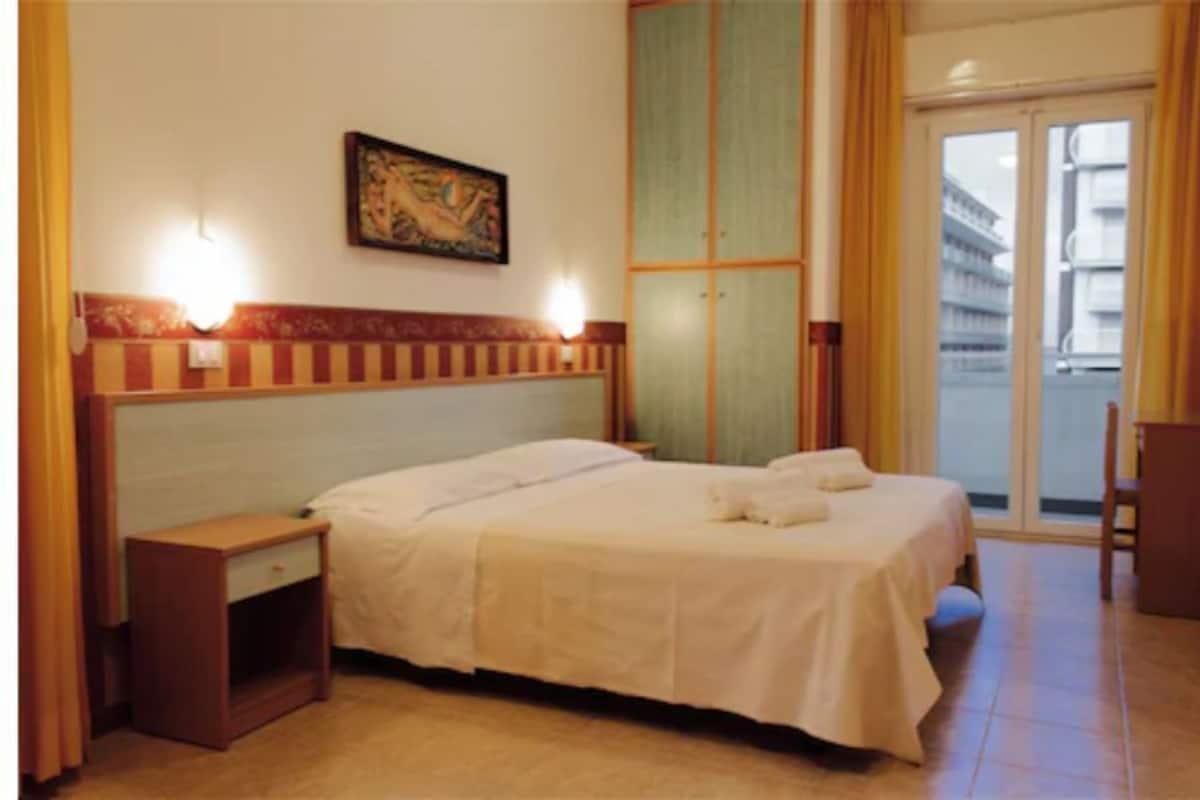 Ein Hotelzimmer mit einem Bett, einem Nachttisch und einem Fenster mit Blick auf Gebäude.