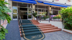 A hotel entrance with a blue awning, a staircase leading to the entrance, and a doormat with a monogram. - Hotel Plaza (Cattolica)
