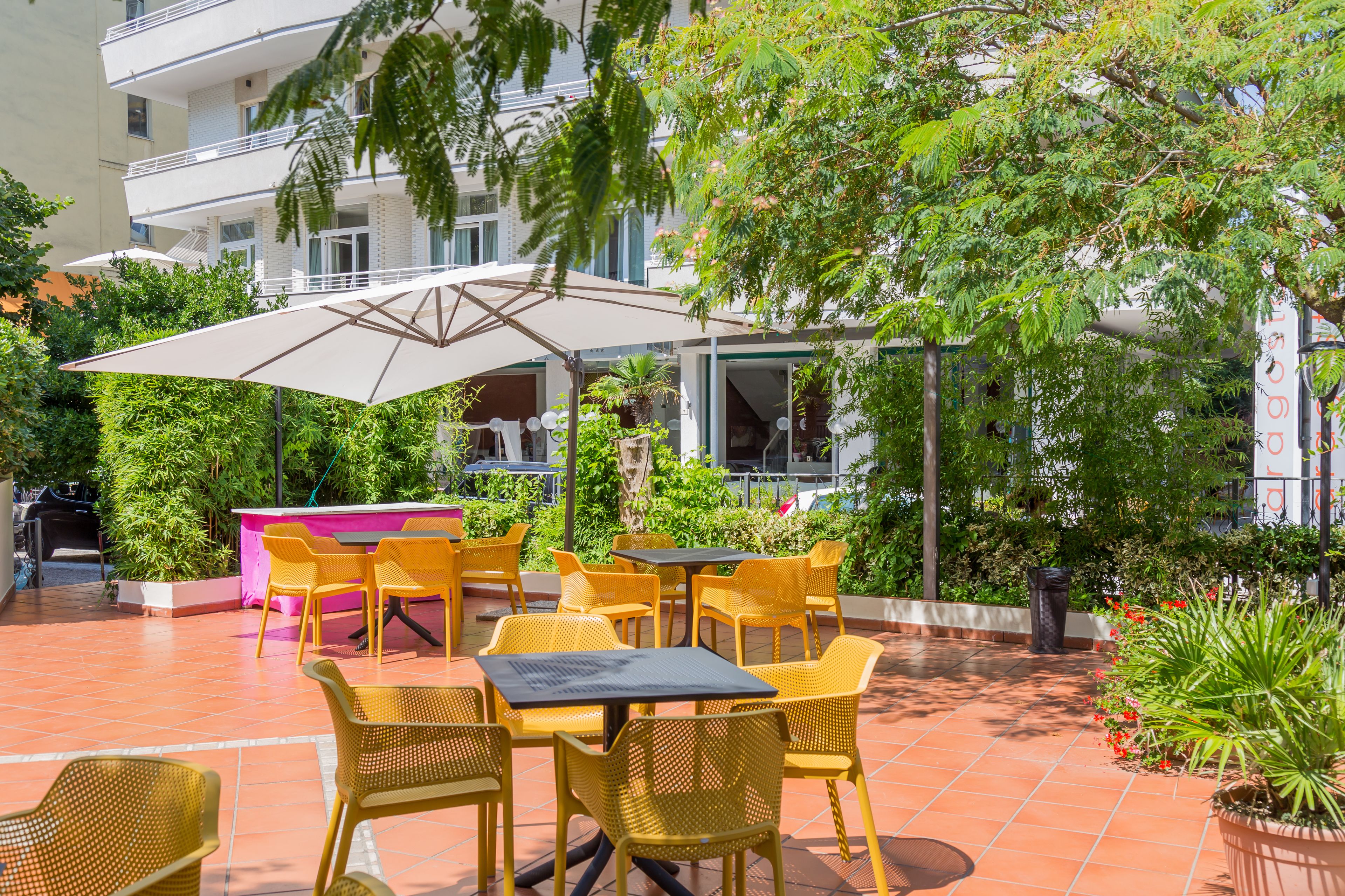 An outdoor patio with yellow chairs and tables, surrounded by greenery and a building in the background.