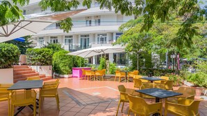 An outdoor patio area with yellow chairs and tables, surrounded by greenery and a building with multiple balconies. - Hotel Plaza (Cattolica)