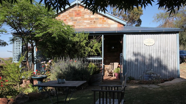 A rustic stone cottage with a metal roof, surrounded by lush greenery and a small patio area with a table and chairs. - Kookaburra Cottage (Burra)