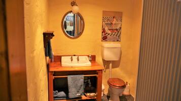 A compact bathroom with a wooden vanity, a round mirror, a toilet, and a small shelf with towels.