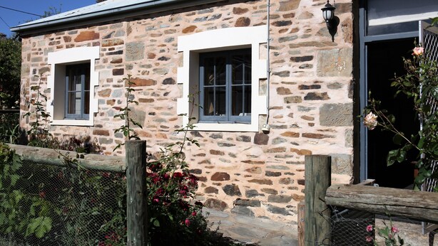 A stone building with two windows, a wooden fence, and a rose bush. - Griffiths Cottage (Burra)