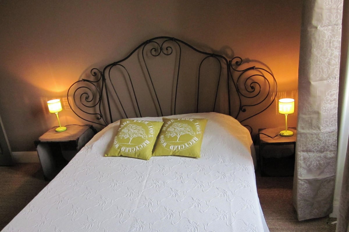 Una cama bien tendida con dos almohadas verdes, un cabecero metálico y dos lámparas de noche.