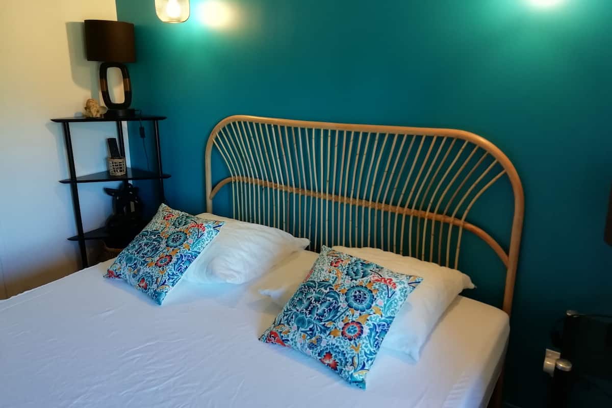 Una cama con colchón blanco y dos almohadas coloridas, cabecero de metal, mesita de noche negra con lámpara y paredes verde azulado.