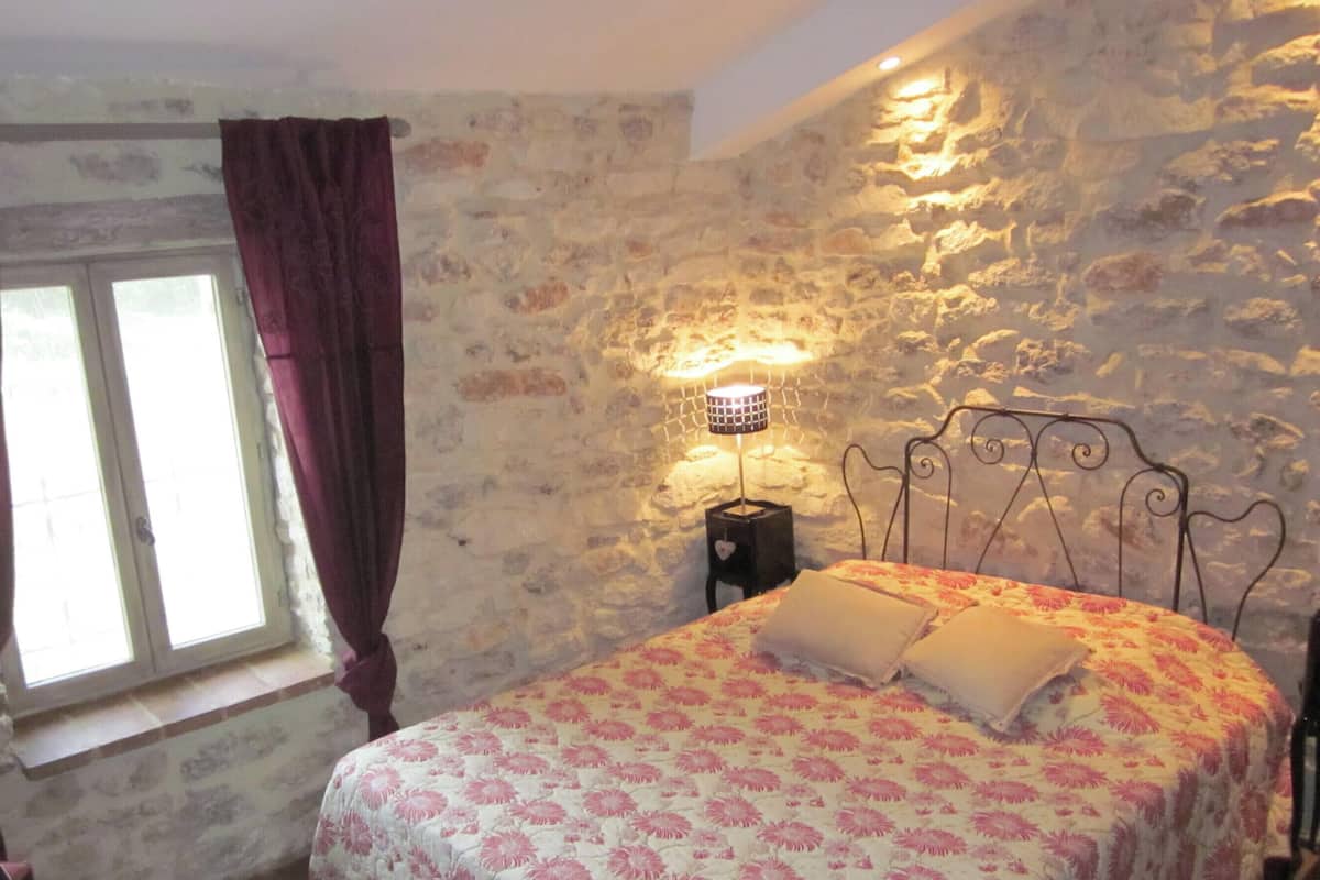 Un dormitorio con pared de piedra, cama con cubrecama floral, mesita de noche con lámpara y ventana con cortinas.