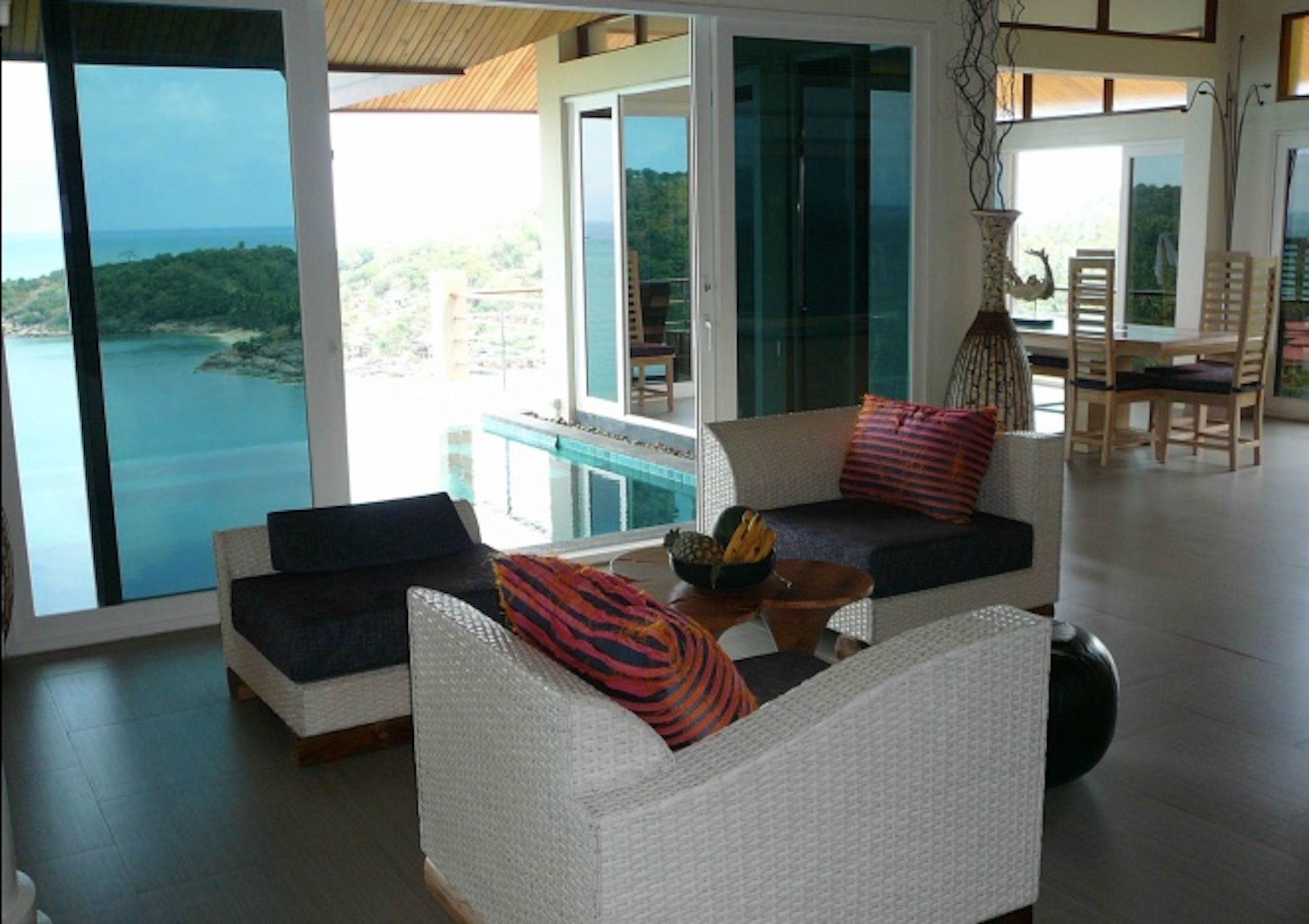 A modern living room with a view of the sea, featuring white wicker furniture, colorful cushions, and a glass door leading to a balcony.