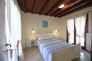 Un dormitorio con cama, dos mesitas de noche, un velador, una ventana con cortinas, un techo con vigas de madera y una lámpara fijada a la pared.