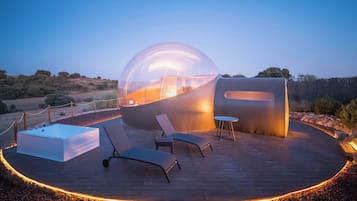 Una estructura moderna con cúpula transparente, un deck de madera, sillones de descanso y una mesa.