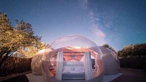 A transparent dome structure with a bed inside, surrounded by a wooden deck and a wooden fence. - Miluna (Hormigos)