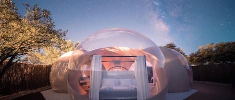 Una estructura de cúpula transparente con una cama en su interior, rodeada por una terraza de madera y una valla de madera.