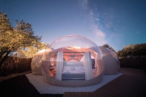Una estructura de cúpula transparente con una cama en su interior, rodeada por una terraza de madera y una valla de madera.