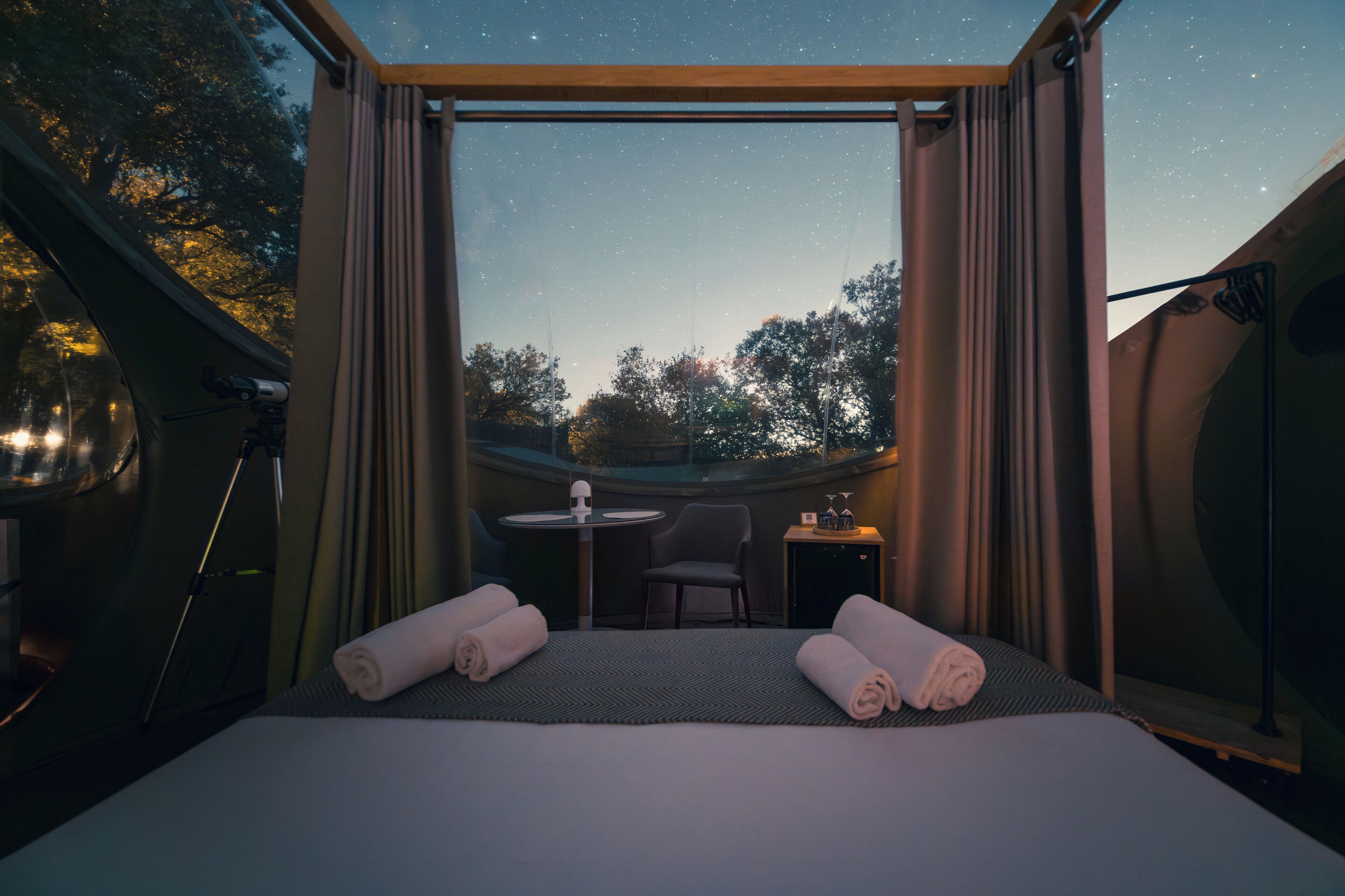 A room with a bed, two rolled towels, a small table, and a chair.