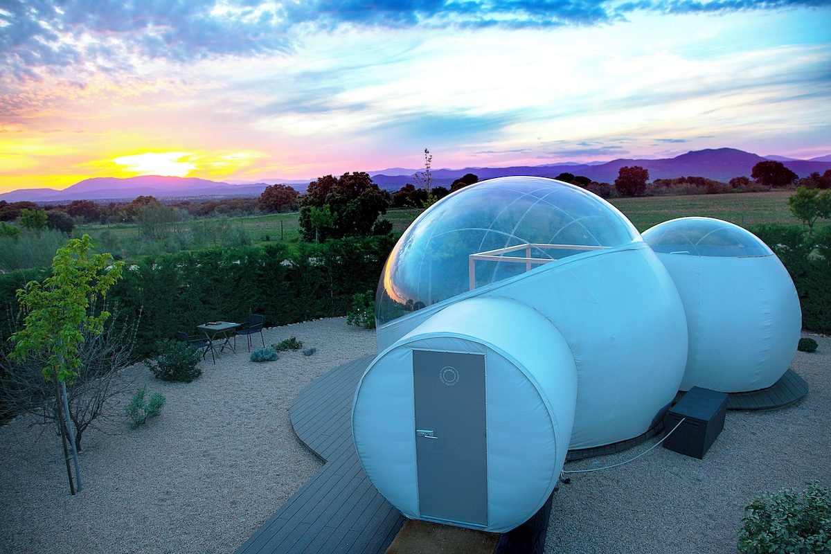 A spherical structure with a door, surrounded by a gravel area and a wooden deck, set against a sunset sky.