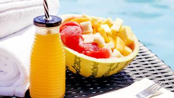 Una mesa de comedor con un bol de fruta, una botella de jugo y una toalla doblada junto a una piscina.