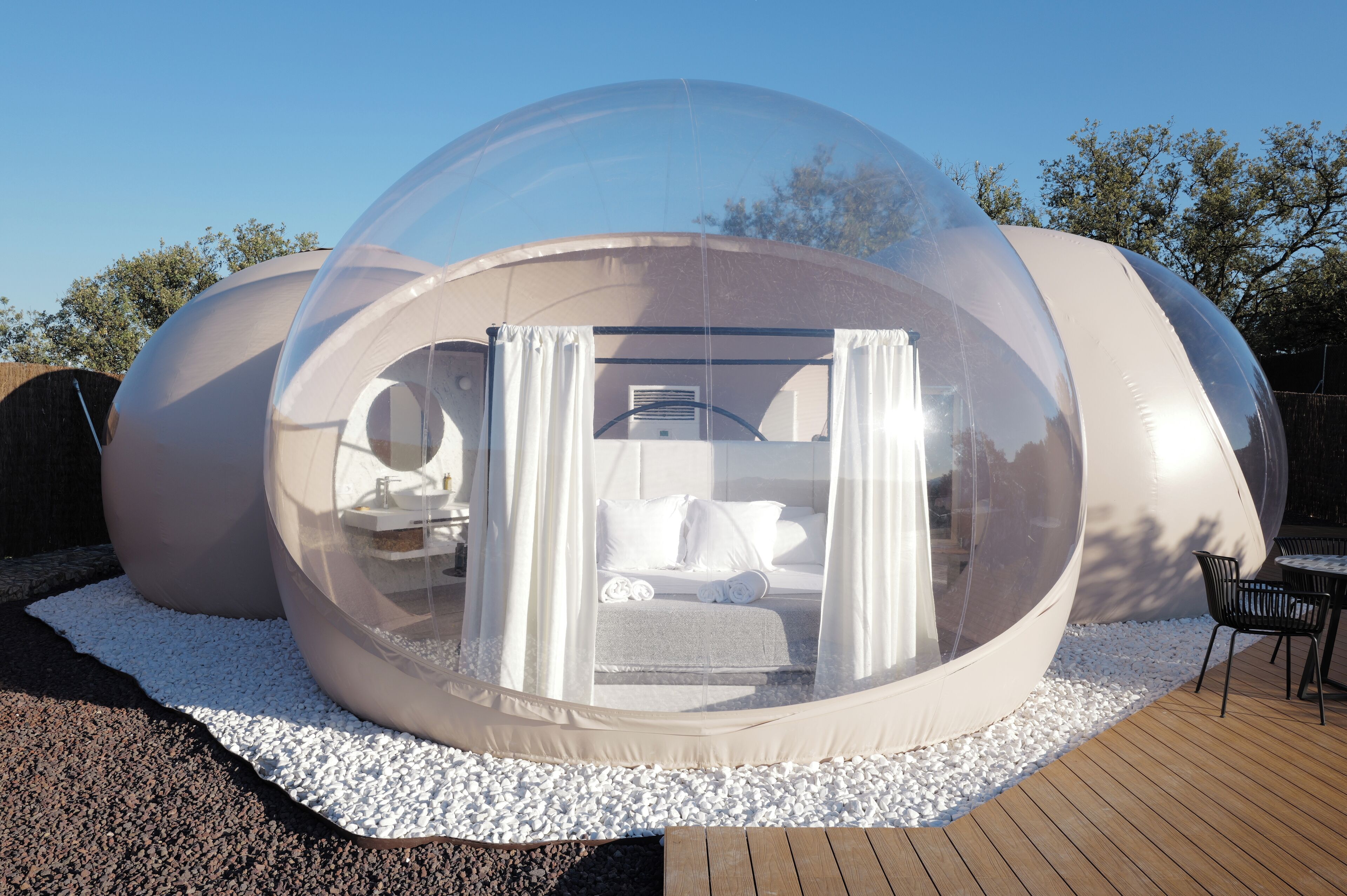 Estructura abovedada transparente con una cama y cortinas, rodeada de guijarros blancos y plataforma de madera.