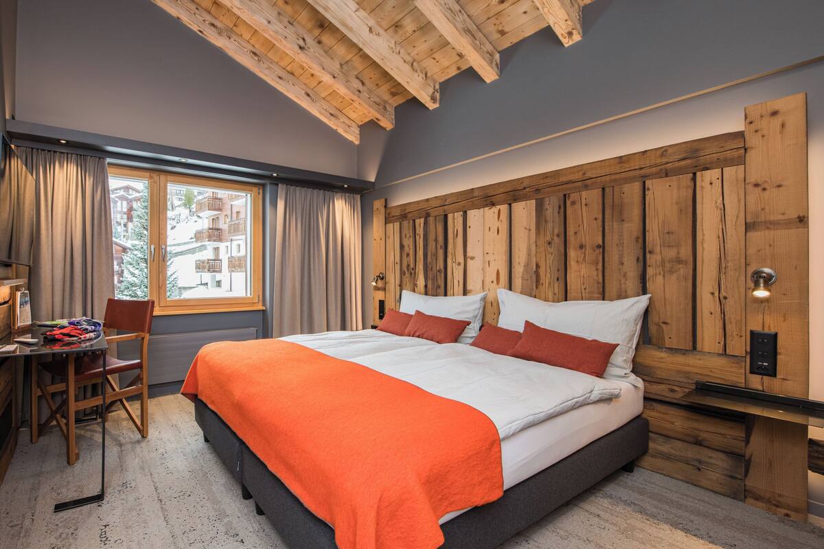 A bedroom with a wooden headboard, a bed with white and orange bedding, a desk with a chair, and a window with curtains.
