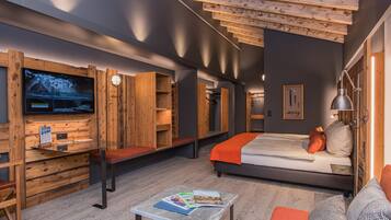 A modern hotel room with a large flat-screen TV, wooden shelving, a bed with orange bedding, and a small table with magazines.