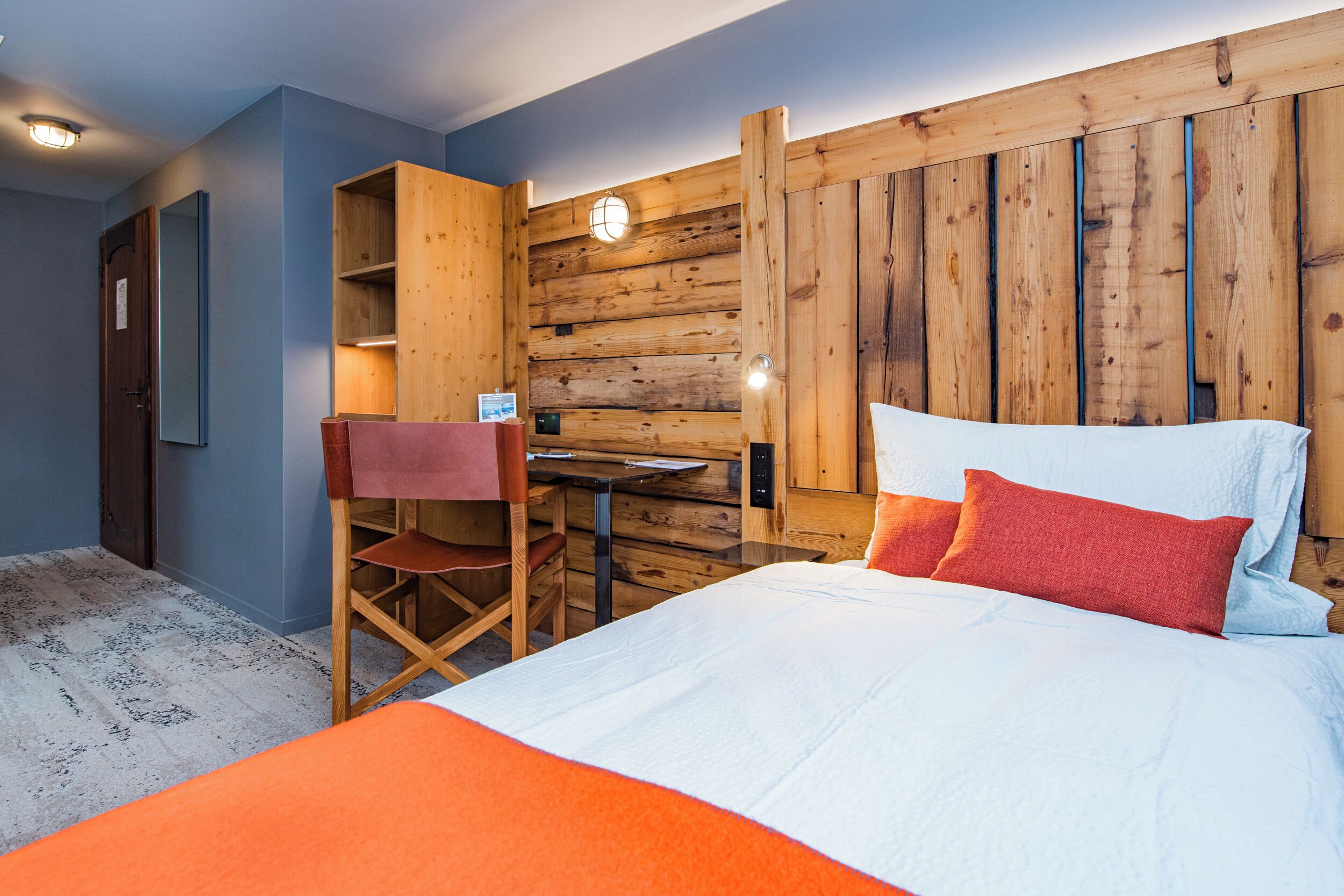 Un dormitorio con cabecera de madera, cama con ropa de cama blanca y roja, un escritorio con silla y un pequeño estante.