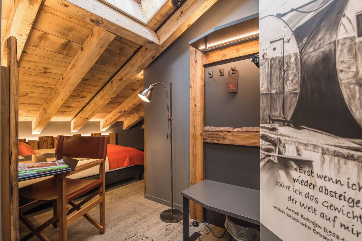 A cozy attic room with wooden beams, a desk with books, a bed with red bedding, and a poster on the wall.