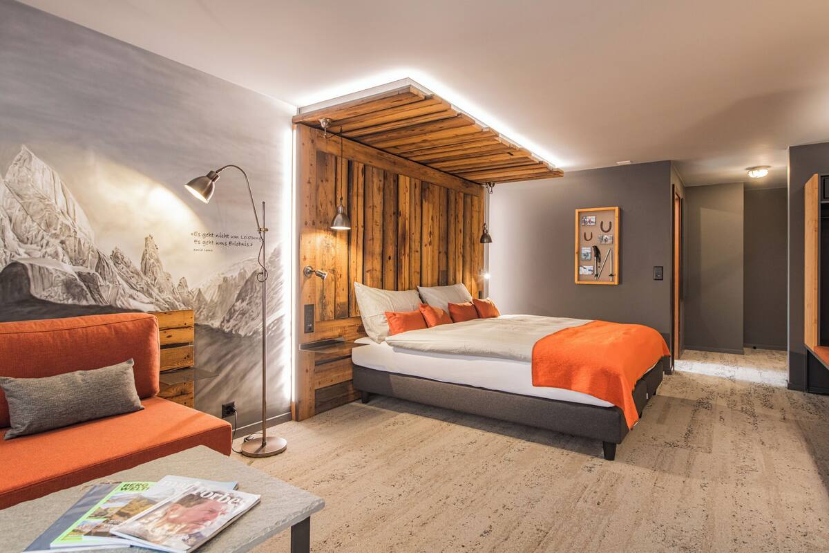 A bedroom with a bed, a sofa, a reading lamp, and a wall with a mountain painting.