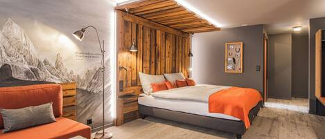 A bedroom with a bed, a sofa, a reading lamp, and a wall with a mountain painting.