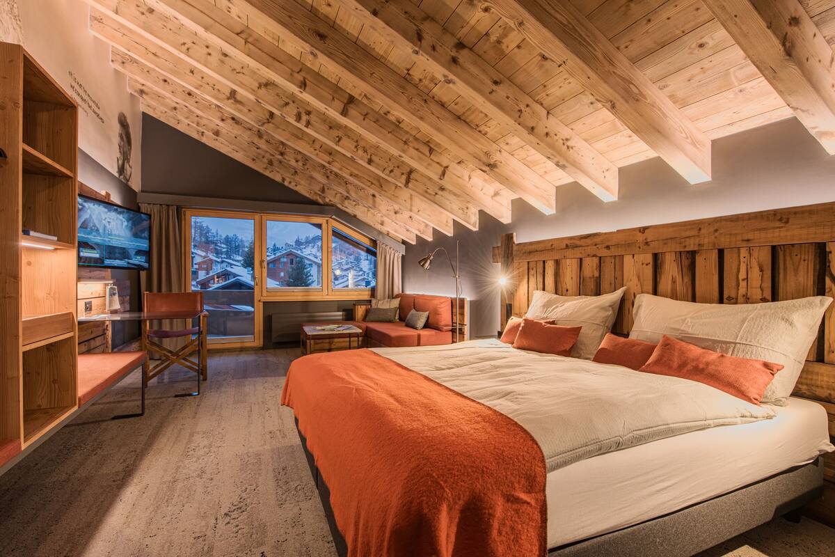 A cozy bedroom with a wooden ceiling, a large bed with orange bedding, a desk with a chair, and a window with a view of snowy mountains.