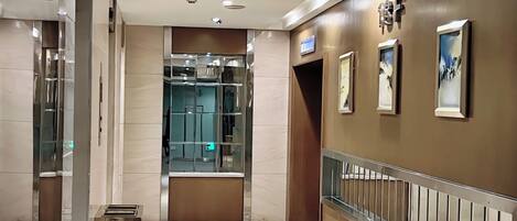 A modern hotel lobby with a marble floor, a glass elevator, and a staircase with a metal railing.