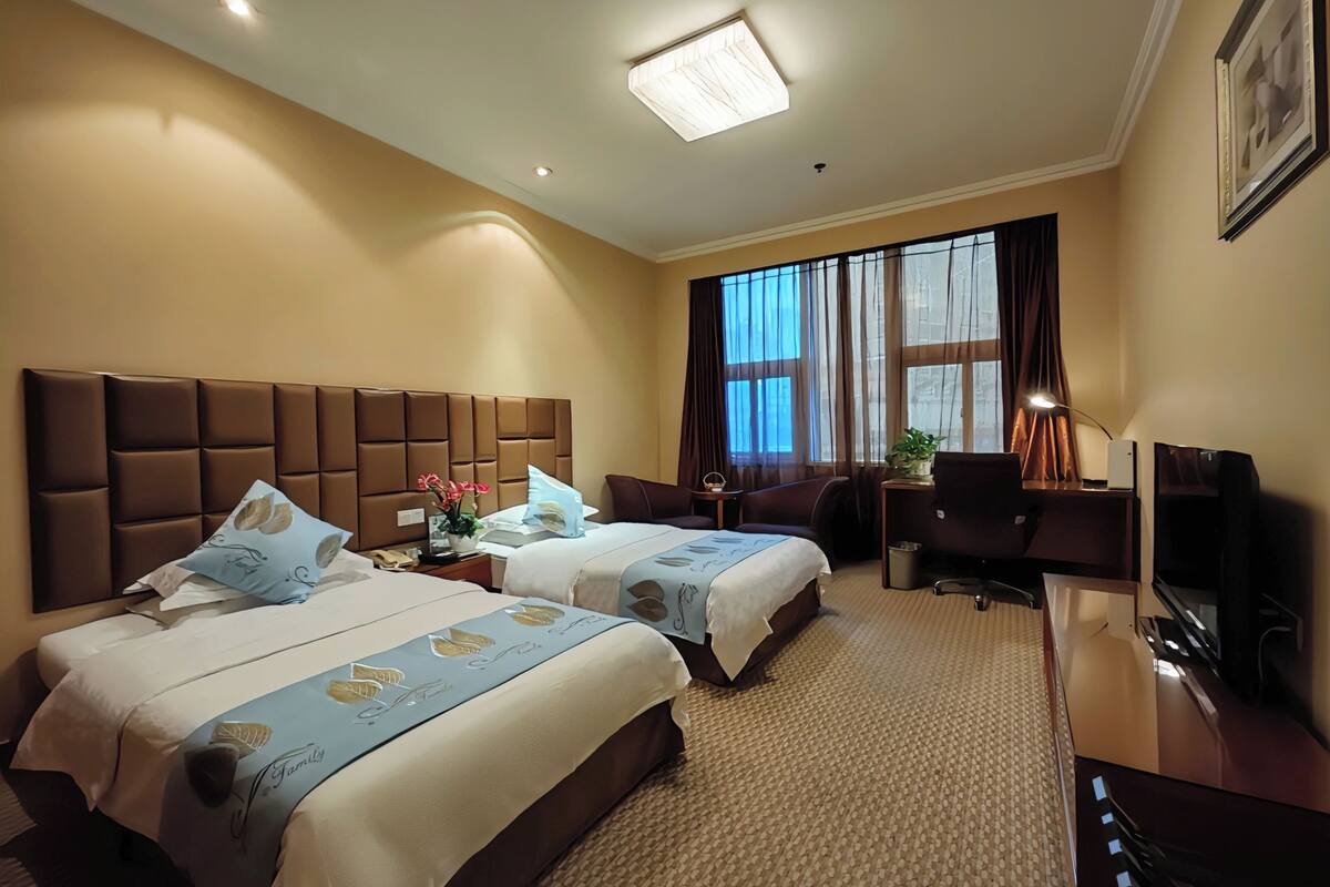 Hotelkamer met twee bedden, een bureau, een stoel, een televisie en een groot raam met gordijnen.
