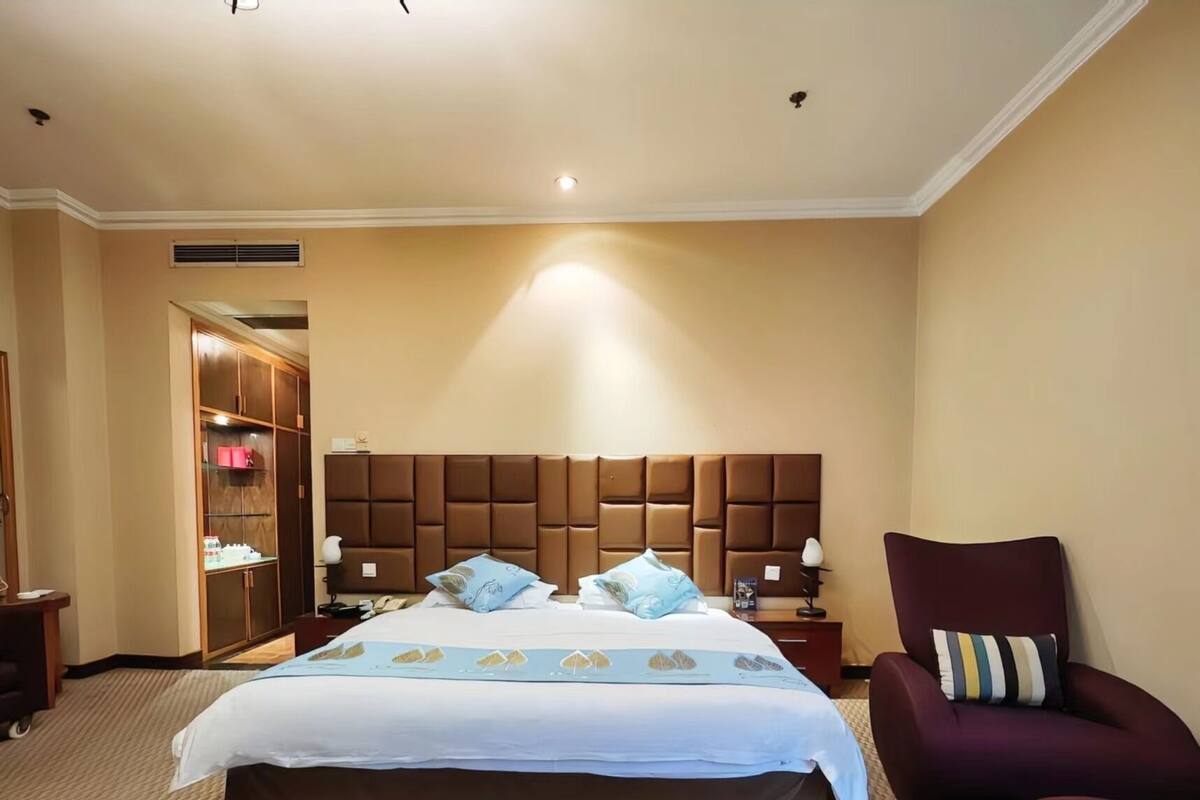 Een hotelkamer met een groot bed, een houten garderobe, een zithoek met een stoel en een klein tafeltje met een lamp.