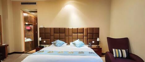 A hotel room with a large bed, a wooden wardrobe, a seating area with a chair, and a small table with a lamp.