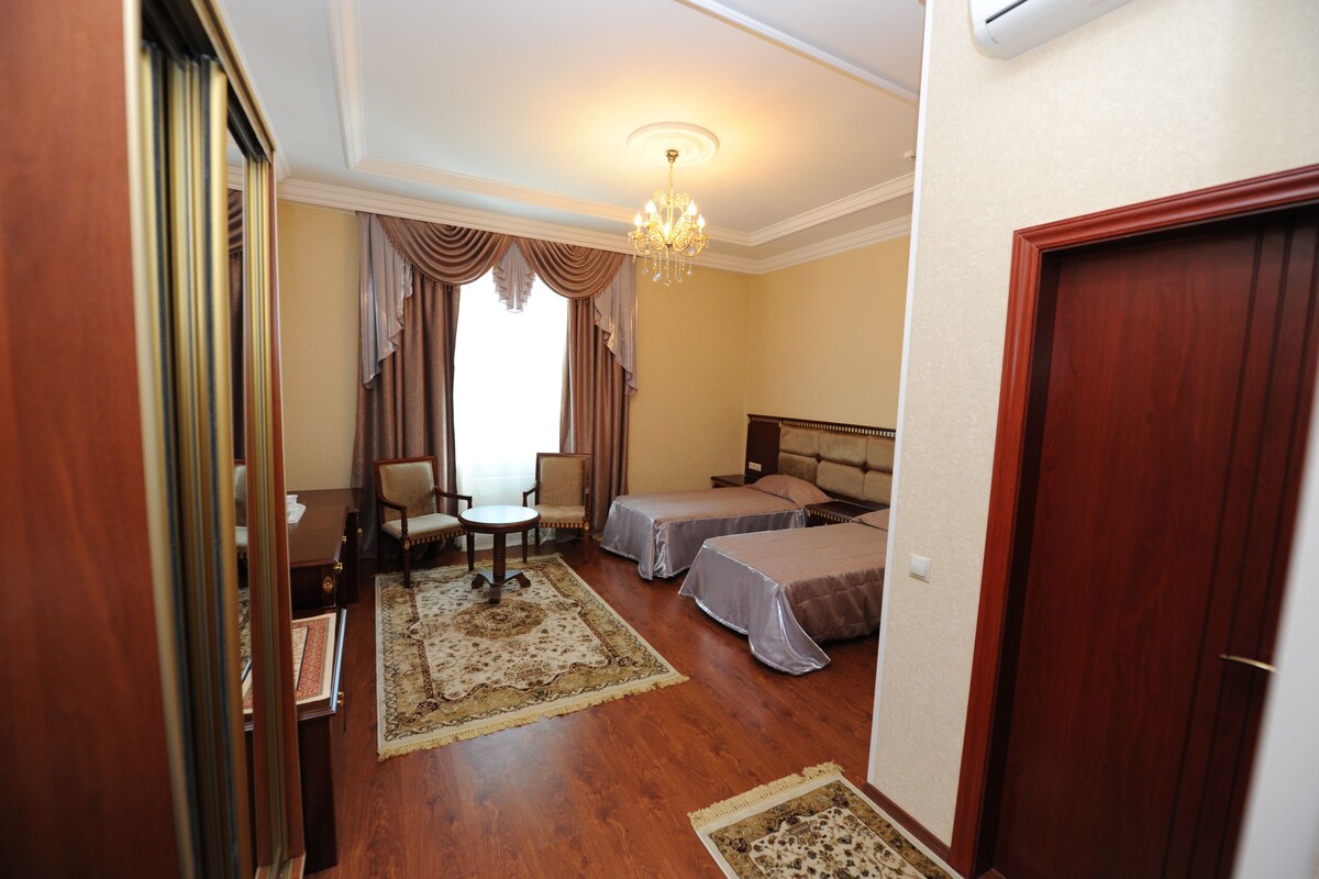 A hotel room with two beds, a chandelier, a wooden door, and a view of the outdoors.