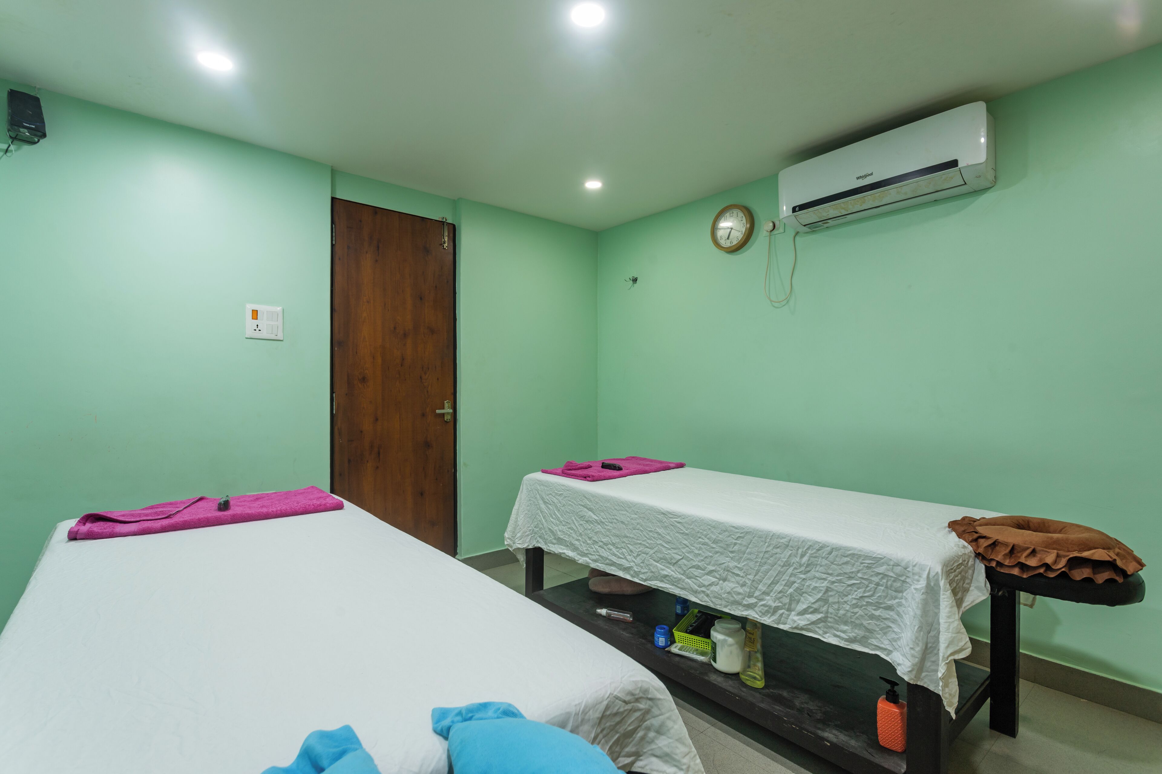 A room with two beds, each with a white bedspread and a pillow. There is a wooden door, an air conditioning unit, and a small shelf with toiletries.