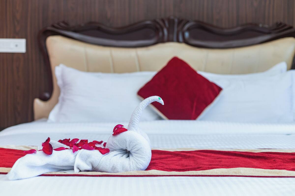 A neatly made bed with a red pillow and a decorative swan made of towels and rose petals.