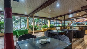 Un restaurant intérieur doté de tables en verre et de chaises en osier, un bar proposant une variété de boissons, et une vue sur les palmiers à l’extérieur.