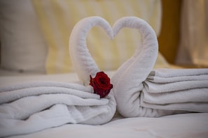 Un lit avec des serviettes blanches soigneusement pliées et disposées en forme de cœur, une rose rouge placée au centre et un oreiller à rayures en arrière-plan.