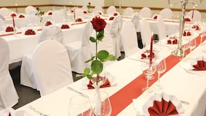Une salle de banquet où les tables sont dressées pour un événement formel, avec des nappes blanches, des serviettes rouges et une rose rouge placée au centre de chaque table.