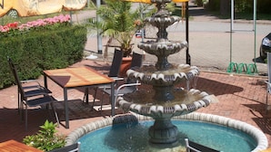 Une fontaine entourée d’un bassin en briques, des tables et des chaises d’extérieur, ainsi que des parasols jaunes.