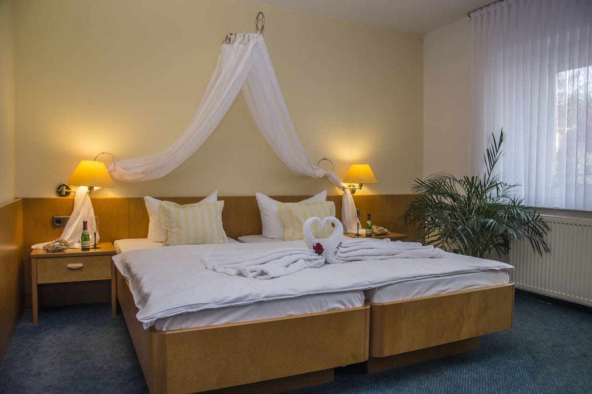 Un lit double avec du linge de lit blanc, deux tables de chevet avec des lampes, une grande plante en pot et un lit à baldaquin.