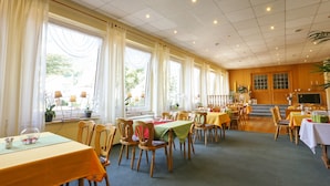 Un espace repas comprenant plusieurs tables et chaises, de grandes fenêtres avec des rideaux et un téléviseur fixé au mur.
