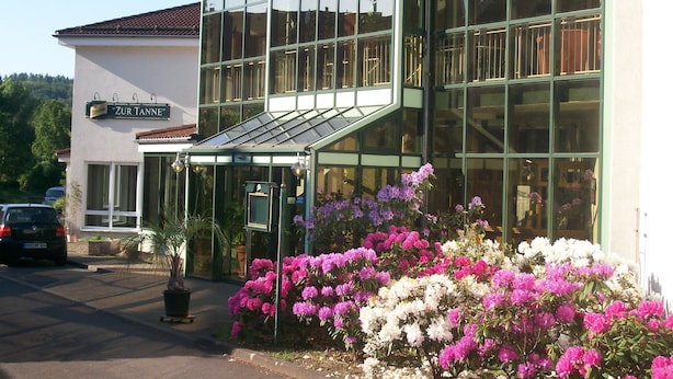 Un bâtiment doté de grandes fenêtres en verre, une enseigne indiquant « Zur Tanne », et une variété de fleurs devant l’entrée.