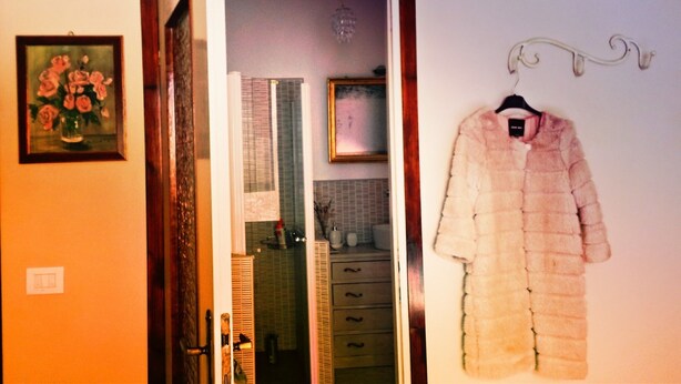 A room with a wooden door, a framed painting, a hanging coat, and a mirror reflecting a tiled wall and a window. - B&B Casa Elena (Gargnano)