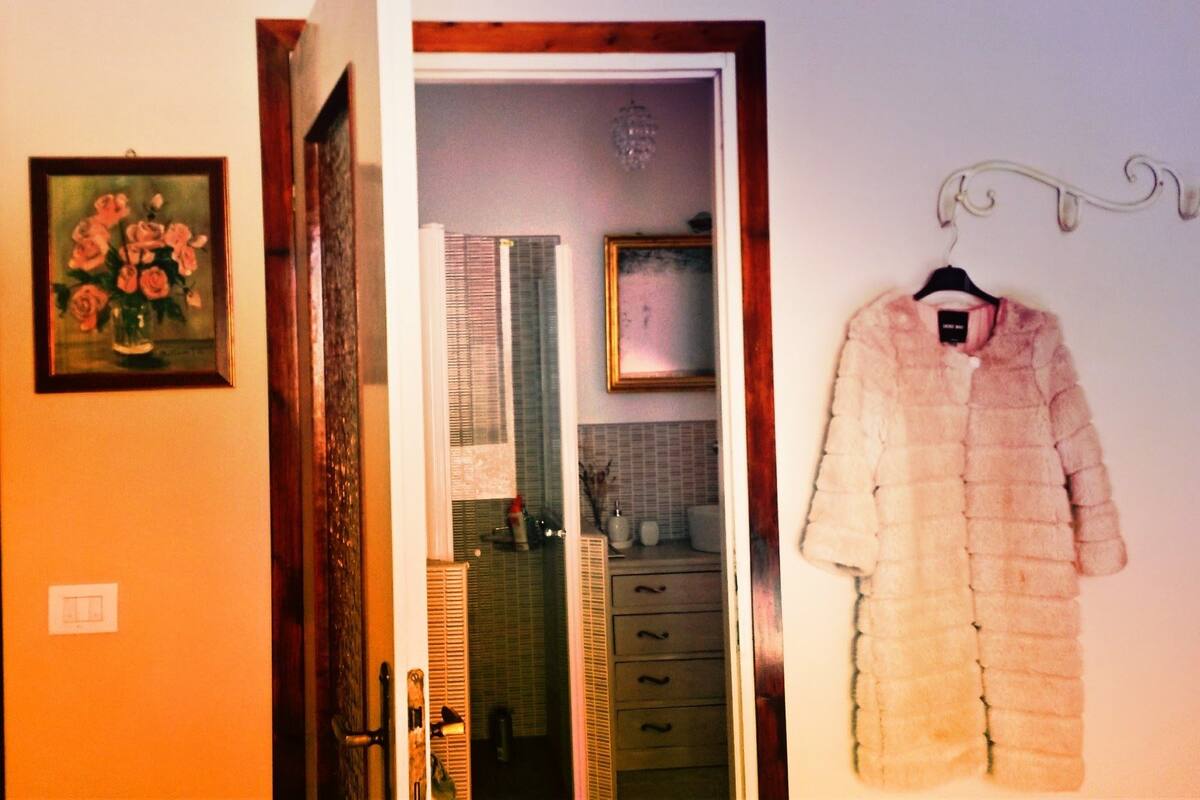 A room with a wooden door, a framed painting, a hanging coat, and a mirror reflecting a tiled wall and a window.