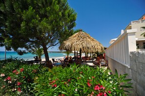 A beachside restaurant with outdoor seating, a thatched umbrella, and a view of the sea.