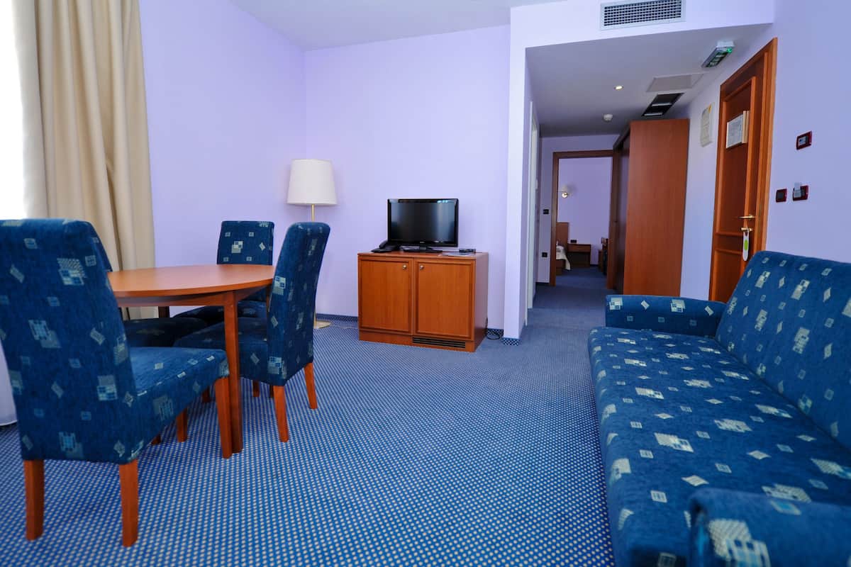 A hotel room with a blue patterned sofa, a wooden TV stand, a dining table with chairs, and a lamp.