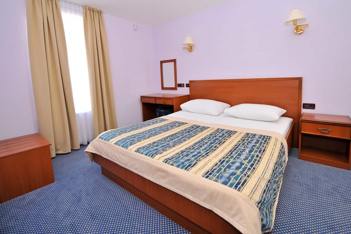 A hotel room with a large bed, a bedside table, a nightstand, a wooden bench, a mirror, and a window with curtains.