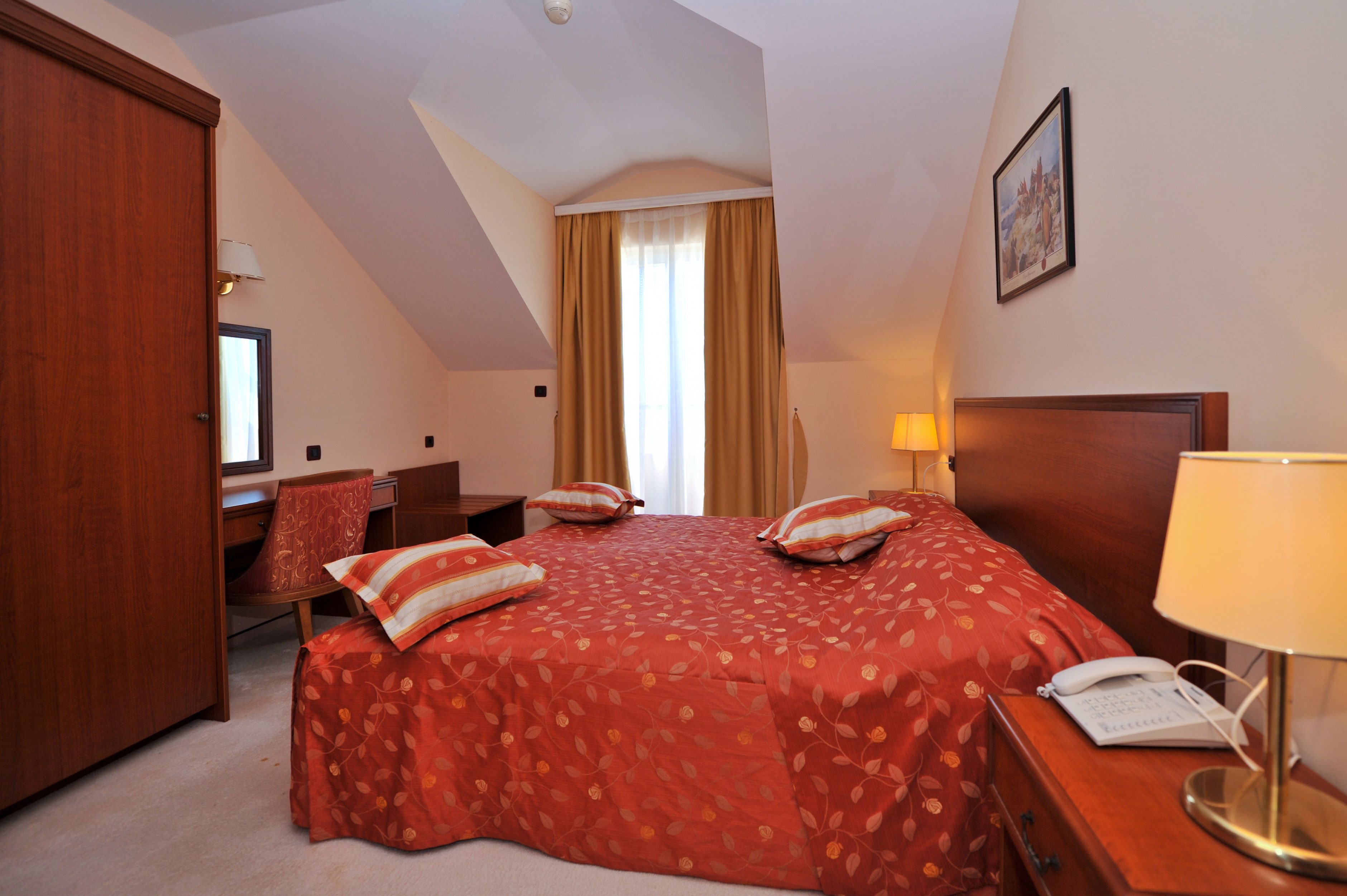 A hotel room with a large bed, a desk, a chair, a wardrobe, a window with curtains, and a lamp.